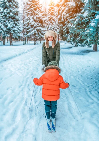 Ibu muda senang dan tertawa, mendorong anaknya anak laki-laki berusia 4-6 tahun, langkah pertama dari ski pada anak-anak. Di musim dingin, di taman kota dengan latar belakang salju dan pohon Natal hijau di salju . — Stok Foto