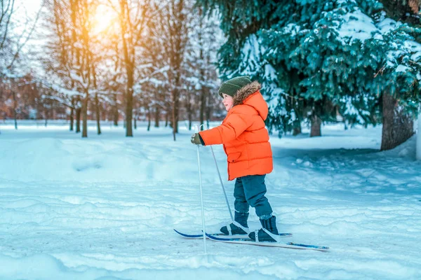 Un niño de 4-6 años con una chaqueta roja en esquís infantiles. En invierno, en el parque de la ciudad, los primeros pasos en esquís, el comienzo de las actividades deportivas desde una edad temprana. Felices caminatas sonrientes delante . — Foto de Stock