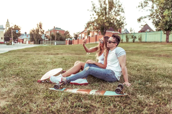 Young couple, selfie photo on phone, video call social networks, online internet application, longboards skate. Happy smiling, concept sporty vacation young family weekends, trendy hipster on grass. — Stock Photo, Image