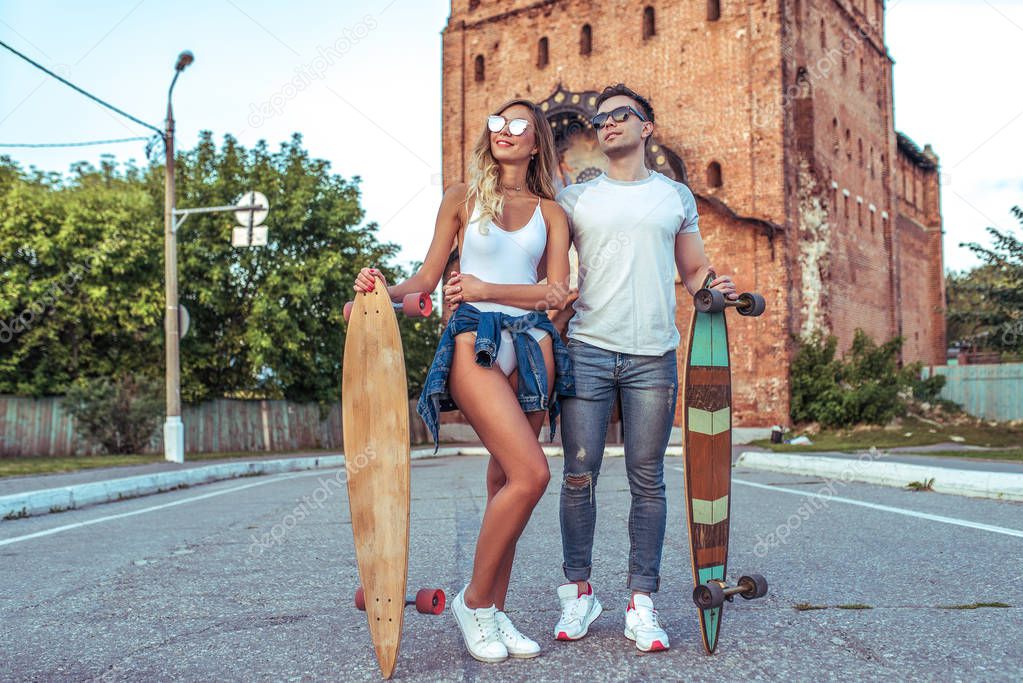 Young couple guy with a girl posing on the road in the hands of longboard skates. Happy smiling and sunglasses, summer in the city. Concept sports vacation young family on weekends, trendy hipsters.