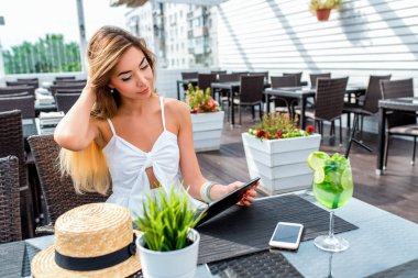Güzel kız beyaz bir elbise bir restoranda bir masada oturan bir yaz kafede. Bir tablet bir film izlerken. İnternette online başvuru sosyal ağlar. Uzun saç düzeltiyor.