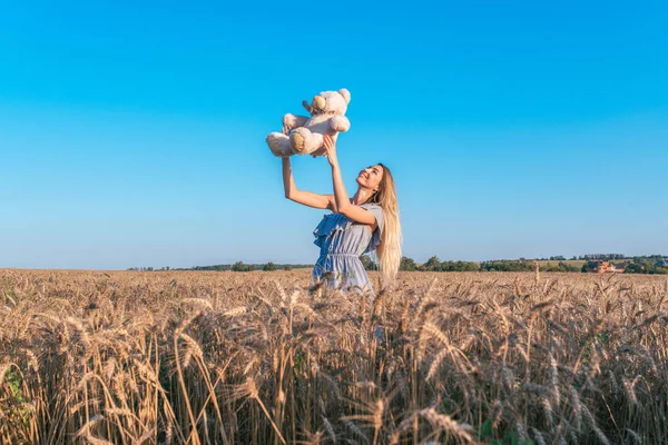Flickan i en klänning, kastar upp en leksak av en nallebjörn. Happy leende sommaren i ett vetefält. Känslor av glädje är gåvan av roliga och friluftsliv. — Stockfoto