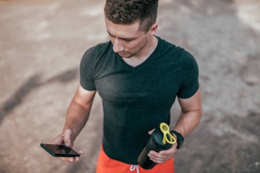 Bir erkek atlet telefon elinde tutuyor, iş için bir uygulama başlattı. Yaz aylarında doğa doğa doğa şişede shaker üst görünümden açıktır. Internet uygulaması, online sosyal ağlar.