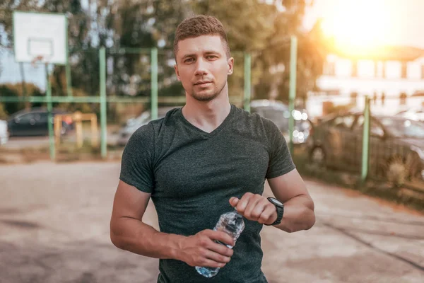Ein männlicher Athlet, im Sommer auf dem Sportplatz, in den Händen eine Flasche Wasser. Durstreduzierung nach dem Sport. Bewegung auf der Straße, frische Luft, aktiver Lebensstil. — Stockfoto