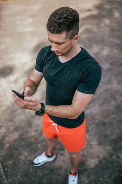 Ein männlicher Athlet hält ein Handy in der Hand, startet eine Anwendung für ein Workout, im Sommer auf Naturblick von oben auf dem Hintergrund von Asphalt. Internet-Anwendung, soziale Netzwerke online. — Stockfoto