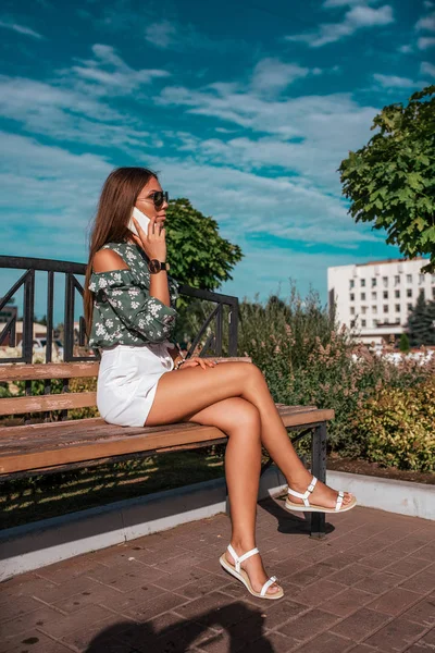 Hermosa chica de verano en el parque se sienta en un banco al aire libre. En las manos del teléfono, llamadas y charlas, en línea, la aplicación de las redes sociales de Internet, falda blanca cielo azul en el fondo . — Foto de Stock