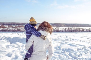 Arka görünümünden, anne kucağında küçük bir çocuk bir çocuğun 3-6 yaşında, şehirde kış öğleden sonra doğada tutar. Metin için özgür yer. Orman sürüklenir ve kar arka planda.