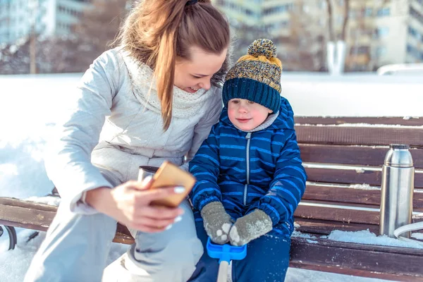 A young mother photographs a 3-5 year old boy on the phone. Happy smiling sit on a bench in the winter in the park. Online selfie application, in the Internet. Thermos mug with tea.