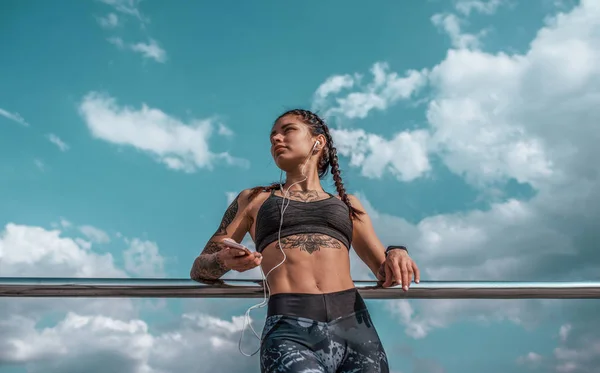 Hermosa chica atlética joven en tatuajes, ropa deportiva, polainas y parte superior. En su mano, el teléfono escucha música en los auriculares. En verano en el descanso de la ciudad después del entrenamiento, entrenamiento de fitness, fondo del cielo . — Foto de Stock