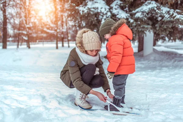 Молодая женщина, мать надевает лыжи зимой в лесном парке, маленькому сыну мальчику 3-6 лет. Заботливая поддержка, первые занятия спортом на улице. Крепление на детских лыжах. Забота о ребенке-родителе . — стоковое фото