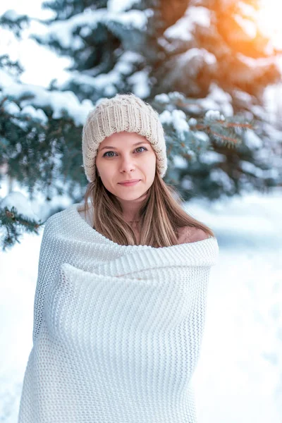 Uma jovem, uma menina bonita com um chapéu branco e um tapete embrulhados, aquece no inverno na floresta no fundo de derivas de neve e árvores de Natal. Feliz sorrindo tempo frio . — Fotografia de Stock