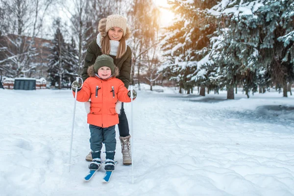 Seorang wanita muda, ibu mendukung dengan hati-hati, bermain ski di musim dingin di taman hutan, seorang anak laki-laki berusia 3-6 tahun. Pelajaran olahraga pertama di jalan. Merawat orang tua-anak. Ruang kosong untuk teks . — Stok Foto