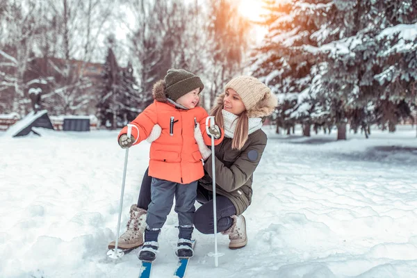Une jeune femme, mère soutient avec soin, petit garçon de 3-6 ans, ski d'hiver dans le parc forestier. Les premières leçons de sport dans la rue. Joyeux sourire jouant avec l'enfant. Espace libre pour le texte . — Photo