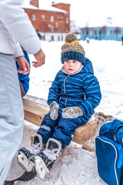 3-5 yıl yaşlı, karla kaplı bir buz pateni pisti üzerinde bir Şehir Parkı içinde belgili tanımlık kış ayakkabı, bir bankta oturan küçük çocuk. Mavi tulum ve siyah paten.