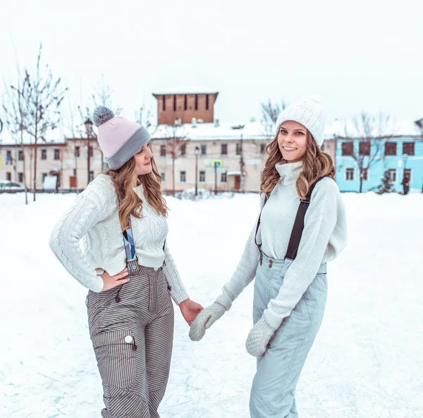 Dua gadis pacar di musim dingin di kota, berdiri di atas baju hangat dan topi hangat, di arena seluncur kota. Happy tersenyum orang berpegangan tangan, santai pada akhir pekan . — Stok Foto