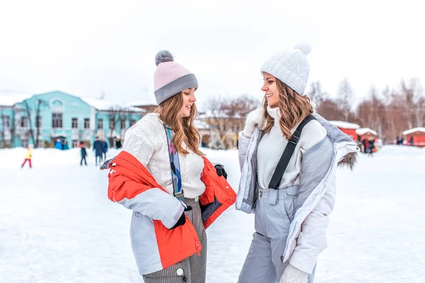 Dwie dziewczyny dziewczyny w zimie w mieście kurtki, stoją w kombinezony, swetry i czapki ciepłe, na lodowisko miasto. Szczęśliwy uśmiechający się ludzie, trzymając się za ręce, relaksując się na weekendy. — Zdjęcie stockowe
