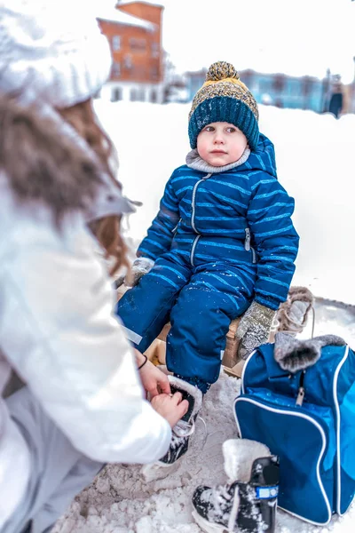 Anya a gyermek s deszkás cipő, a 3-5 éves kisfiú cipő, ül egy padon a cipő, a tél, a hóval fedett jégpálya a Városligetben. Kék overall és fekete görkorcsolya. — Stock Fotó