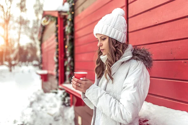 Piękna dziewczyna w zimie w mieście w pobliżu czerwony okiennice, filiżance kawy jest ocieplenie i dmuchanie gorącej herbaty w jej ręce. Wolnego miejsca. Biały kapelusz i ciepły zimowy płaszcz. — Zdjęcie stockowe