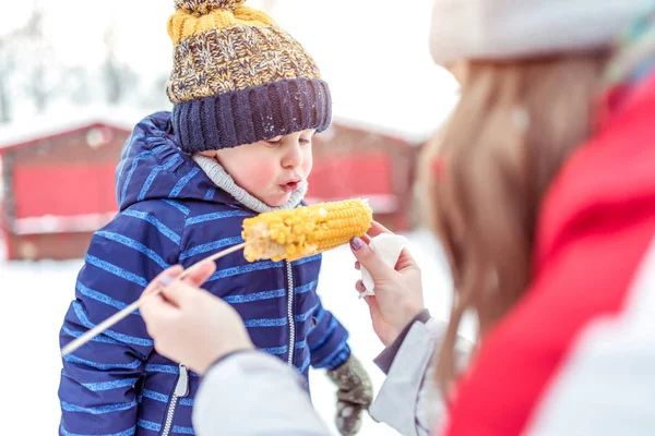 Женщина, мать кормит 3-5-летнего мальчика руками своего сына зимой на выходных. Вареная кукуруза на палочке. Синий комбинезон теплая шляпа . — стоковое фото