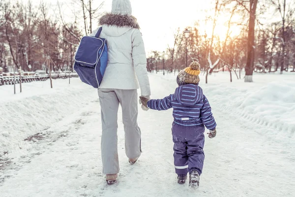 Ibu dengan anaknya kembali ke rumah setelah berjalan-jalan di taman di musim dingin di tengah salju dan salju melayang. Memegang anak kecil dengan tangan . — Stok Foto