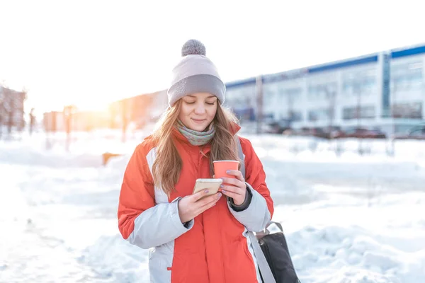 A girl with a phone stands in winter in city, a warm hat and a winter jacket. In his hand a phone, writes and reads a message on the Internet. Online in social networks, a smartphone application.