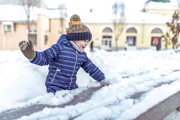 Ein kleiner Junge im Alter von 3-6 Jahren spielt im Winter in der Stadt, hat Spaß beim Schneeballspiel, sammelt Schnee von der Bank, Emotionen der Freude und Spaß am Wochenende in der Natur im Winter. — Stockfoto