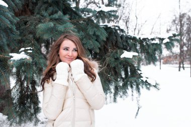 Bir esmer kız karlı Noel ağaçları Park kış arka plan karşı ayakta. Mutlu bir gülümseyen kadın rüyalar, metin için özgür yer. Sıcak beyaz eldivenler ve ceket sıcak, kar sürüklenir arka planda.