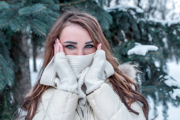 Girl in winter in a city park on background of snow and Christmas trees. In winter in forest, close-up look eyes. She covers her face with a white scarf, emotions of tenderness, warms up cold day. — Stock Photo, Image