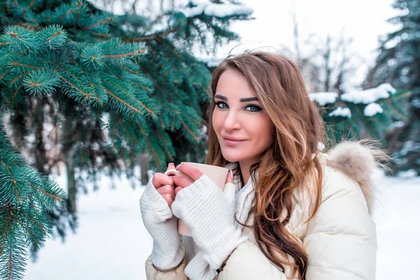 Flicka på vintern i en park på bakgrund av snö och julgranar. På vintern i skogen, närbild titta ögon. I handen håller en mugg av dryck, varmt te, kaffe. Ledigt utrymme för text. — Stockfoto