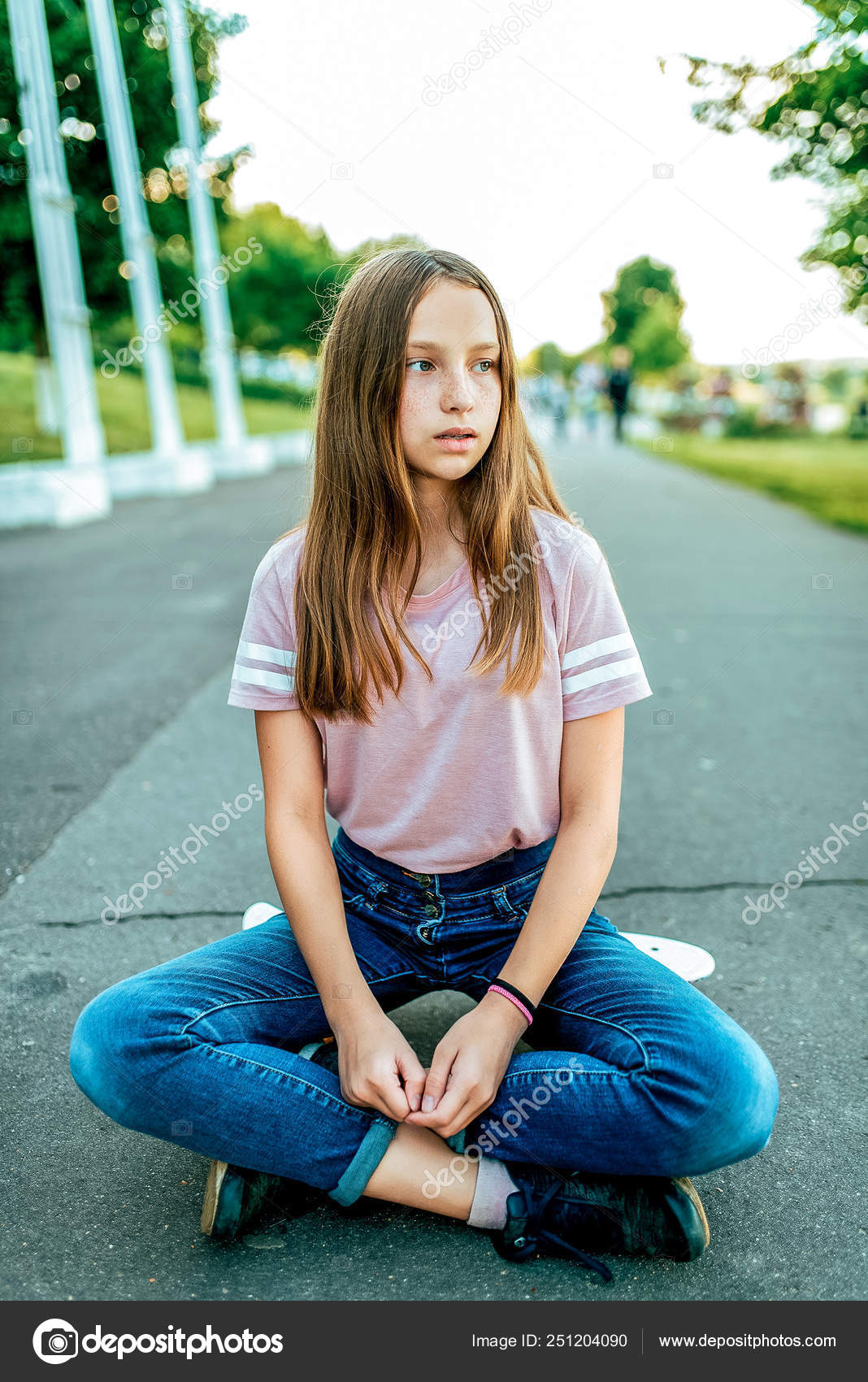 школьница 12 лет Маленькая девочка школьница 12-16 лет, на улице сидит скейтборд летом в  парке. Отдых после прогулки и катания на коньках на борту. Сидят в позе  лотоса, джинсы и повседневная одежда в футболке . —