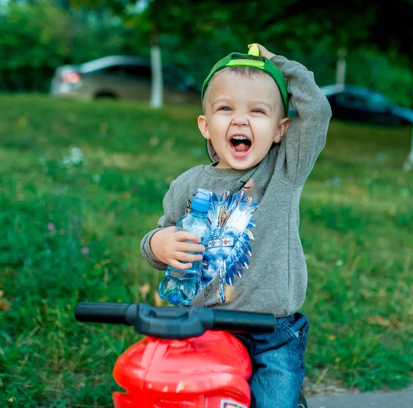 Anak kecil berusia 3-5 tahun, duduk di atas mainan. Di musim panas di taman kota. Di tangannya sebotol air, berteriak, emosional gembira, bersenang-senang, beristirahat di luar akhir pekan . — Stok Foto