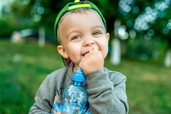 Liten pojke 3-5 år, närbild. I sommaren i stadsparken. I sin hand en flaska vatten vilande lyckligt leende, känslomässigt joyful, ha roligt, på en helg utanför. — Stockfoto