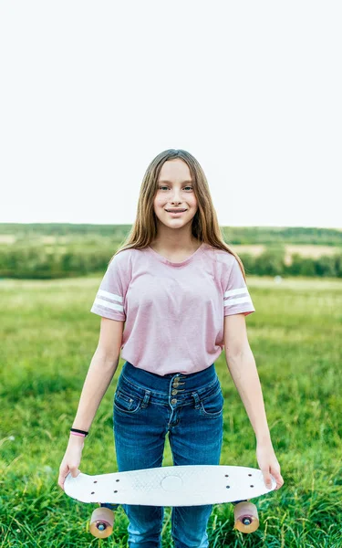 Kleines Mädchen im Alter von 12-16 Jahren, im Sommer auf der Straße im Park. in den Händen eines Skateboards, glücklich lächelndes, emotional freudiges Mädchen. — Stockfoto