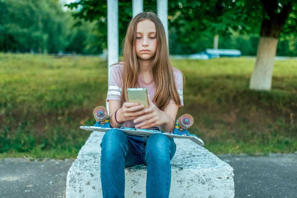 Školačka malá holčička 12-16 let, sedí venku v létě v ulici. Drží, skateboard, telefon, online aplikace, internetové sociální sítě ve svých rukou, čte a zapisuje zprávu v konverzaci. — Stock fotografie