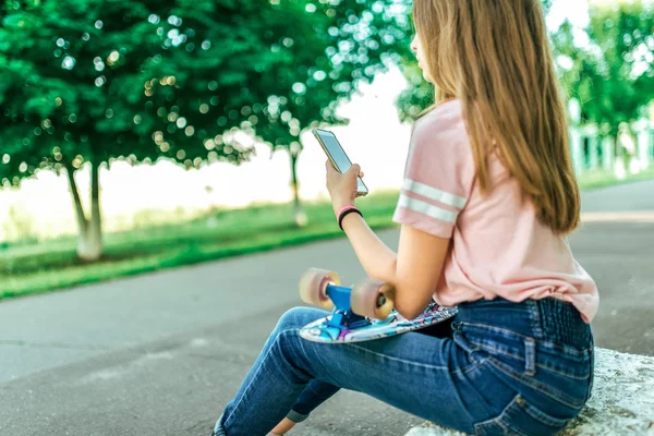 Niña colegiala de 12-16 años, se sienta afuera en el parque de verano. Espacio libre. Sostiene monopatín, teléfono, aplicación en línea, redes sociales de Internet, lee y escribe mensajes en el chat . —  Fotos de Stock