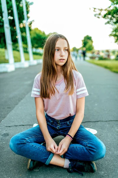 Una niña colegiala de 12-16 años, en la calle se sienta monopatín en verano en el parque. Descansando después de caminar y patinar a bordo. Se sienta en posición de loto, jeans y ropa casual en una camiseta . —  Fotos de Stock