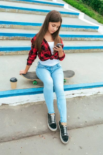 Gadis sekolah 13-16 tahun duduk di tangga skateboard di telepon genggamnya, secangkir teh kopi. Aplikasi obrolan daring di jaringan sosial, cetak dan menonton video di smartphone Anda . — Stok Foto
