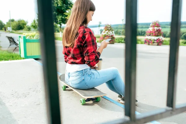 Girl schoolgirl 13-16 years old sits on skateboard stairs in her hand phone. Online chat application in social networks, prints and watches videos on your smartphone. Railing fence on the background. — Stock Photo, Image