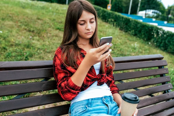 Ragazza studentessa 13-16 anni seduta su una panchina nel suo telefono. Applicazione di chat online sui social network, stampa e guarda video sul tuo smartphone. Una tazza di tè al caffè in estate in città . — Foto Stock