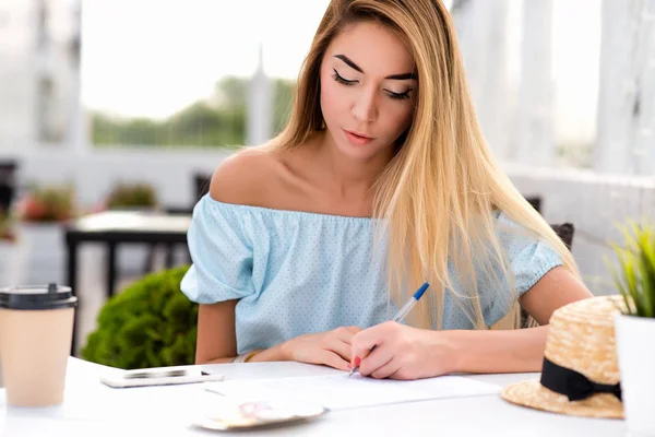 Piękna dziewczyna z długimi włosami, w lato café restaurant, podpisała kontrakt papieru, umowy, stawia malarstwo w pióro. Biznes pani w kawiarni, wypełnia dokument. Telefon, filiżankę kawy i herbaty na stole. — Zdjęcie stockowe