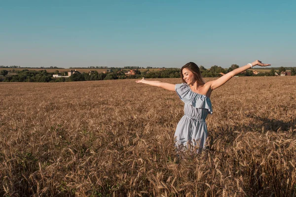 En vacker och ung flicka står i en sommar vetefält. Armarna utsträckta glada leenden jublar Soluppgång och solnedgång. Ledigt utrymme för text. Känslor av lycka ömhet i en blå klänning. — Stockfoto