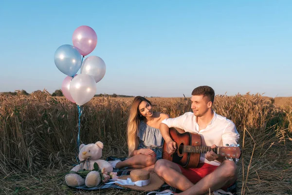 Ein junges Paar sitzt auf karierten Sommerkleidern in einem Weizenfeld. Entspannung in der Natur des Gitarrespielens. romantische Beziehung zwischen Mädchen und Mann. Ballonfarbene Kugeln, Spielzeug Babybär. — Stockfoto