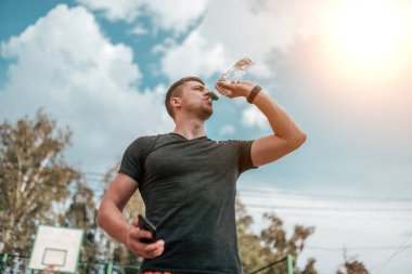 Erkek atlet runner, dinlenme ve bir su şişesi içme suyu. Elinde, telefon denetimleri nabız, uygulamayı çevrimiçi smartphone üzerinde. Park spor alanına yazın. Egzersiz yaşam tarzı.