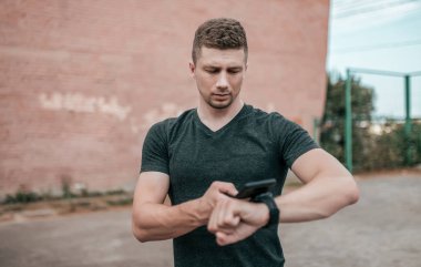 Bir erkek atlet yaz spor şehirde zemin üzerinde duruyor. Bir akıllı telefon elinde, nabız saati denetler. Bilezik verileri eşitler online başvuru. Egzersiz açık hava yaşam tarzı.