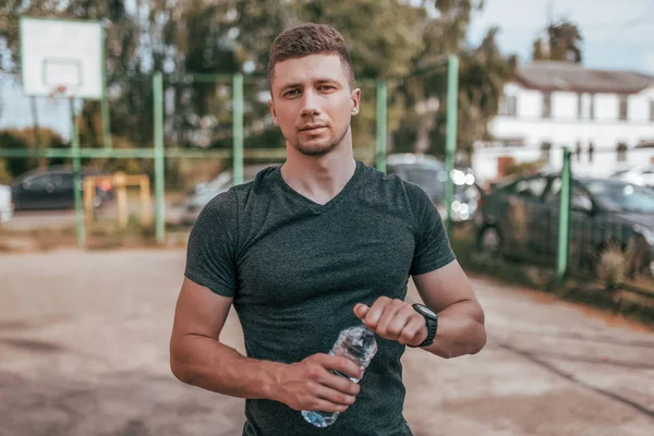 Ein männlicher Athlet steht im Sommer auf einem Sportplatz in der Stadt. in seiner Hand eine Flasche Wasser, die sich nach dem Sport ausruht. Trainer-Typ im Armband der Armbanduhr nach dem Joggen. Workout im Freien. — Stockfoto