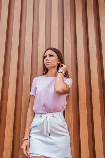 Een mooi meisje in de zomer in de stad, staat tegen een gestreepte houten muur. Ziet er in de verte, rechtzetten haar haar, een roze T-shirt, een witte rok. — Stockfoto
