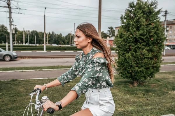 Gadis di musim panas di kota, naik sepeda, dengan latar belakang pohon dan mobil hijau. Dalam sweter hijau dan rok putih . — Stok Foto