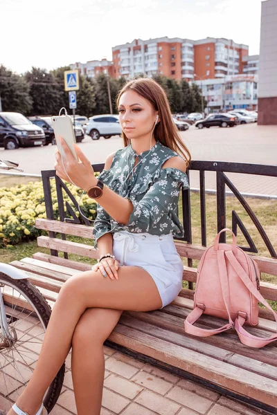Mujer en el verano en la ciudad, se sienta en un banco en su teléfono inteligente de la mano, lee las redes sociales de chat, toma fotos, escucha música en los auriculares . — Foto de Stock