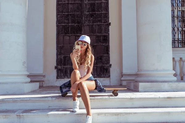 Sports girl with skateboard, longboard summer in city. Fashionable woman in her hand telephone, communication Internet, social networks chat, glasses and a baseball cap, tanned skin and slim figure.