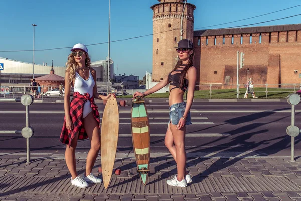 Dua gadis gadis kota musim panas, siswa skateboard modis, pakaian santai longboard. Berdiri di persimpangan berpose. Background jalan mobil dan trotoar, ruang kosong . — Stok Foto